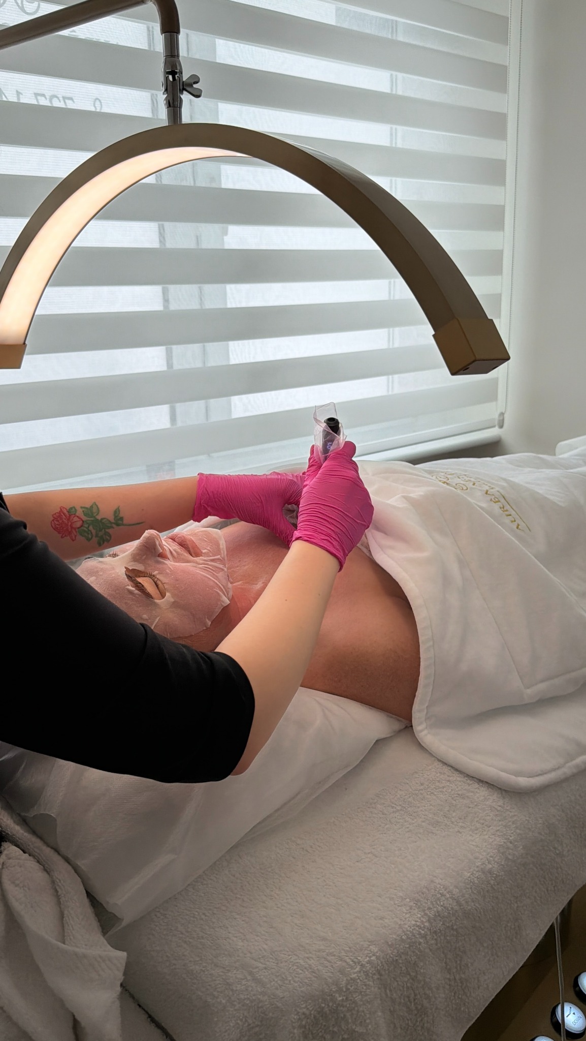 Klientka z maską na twarzy leży na łóżku kosmetycznym, podczas gdy kosmetyczka w różowych rękawiczkach wykonuje zabieg. Widoczna lampa kosmetyczna i tatuaż na ramieniu.