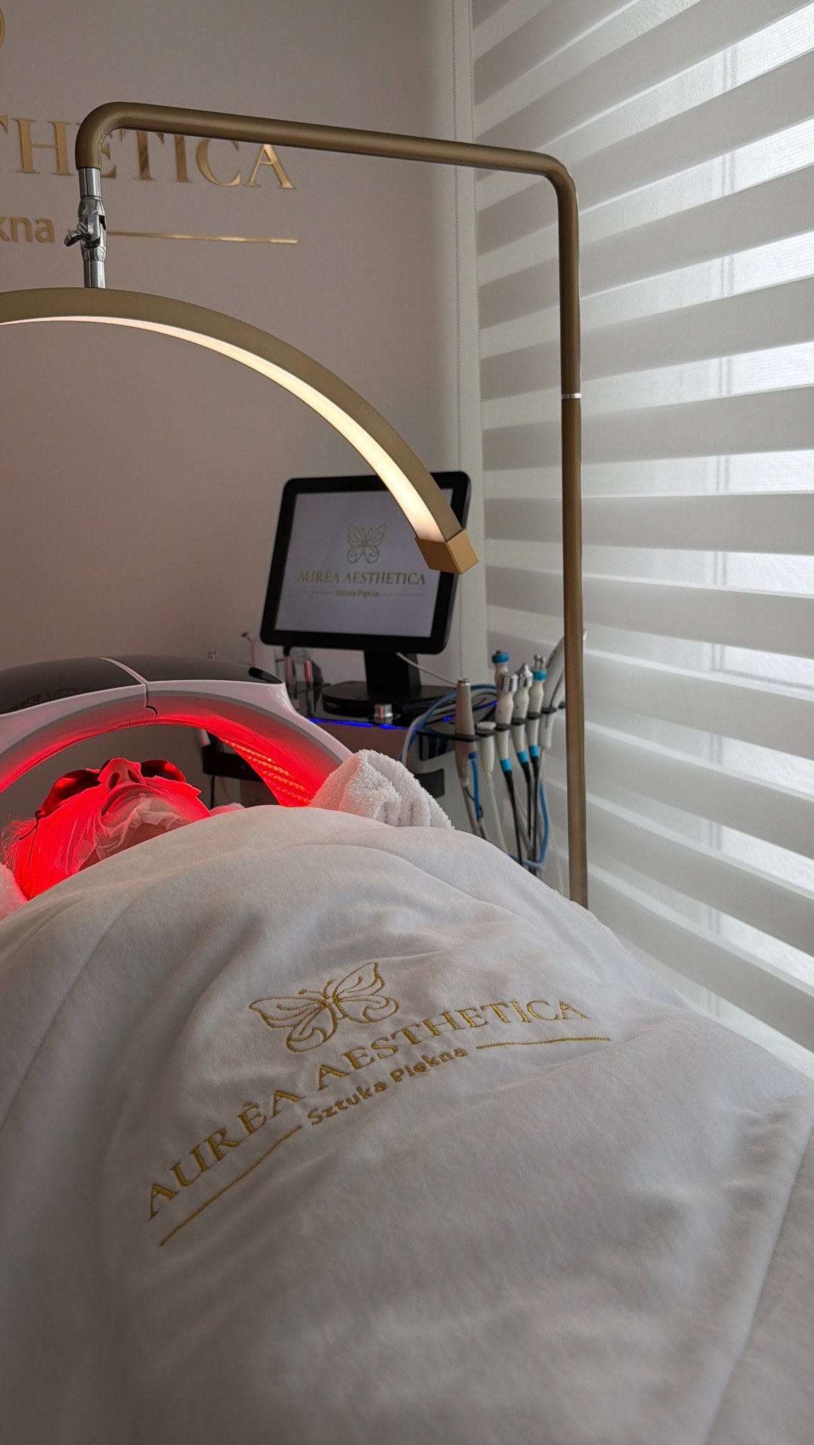 Klientka poddaje się zabiegowi na ciało w salonie kosmetycznym, oświetlona lampą LED. Widoczne narzędzia i monitor z logo salonu Aurea Aesthetica.