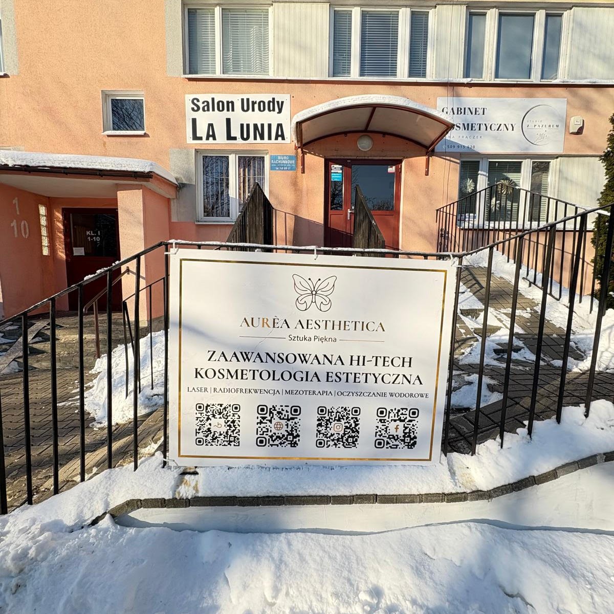 Wejście do salonu urody 'La Lunia' z szyldem 'Aurea Aesthetica' oferującego zaawansowaną kosmetologię estetyczną, w zimowej scenerii.
