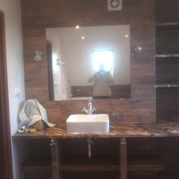 Usługi Remontowo-Budowlane Robert Stefaniak - Nowoczesna łazienka z drewnopodobnymi płytkami, białą umywalką i lustrem, w którym odbija się osoba robiąca zdjęcie. Widoczne półki i akcesoria łazienkowe.