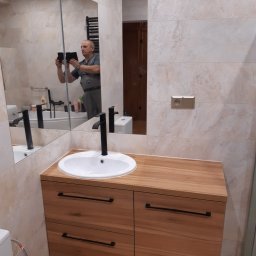 Usługi Remontowo-Budowlane Robert Stefaniak - Nowoczesna łazienka z drewnianą szafką pod umywalką i beżowymi płytkami. W lustrze odbija się osoba robiąca zdjęcie telefonem. Widoczna armatura i akcesoria łazienkowe.