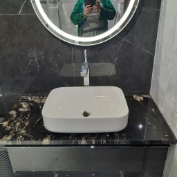 Usługi Remontowo-Budowlane Robert Stefaniak - Nowoczesna łazienka z umywalką nablatową na blacie z czarnego marmuru, podświetlane lustro okrągłe. W odbiciu widoczna osoba robiąca zdjęcie.