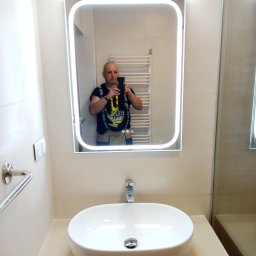 Usługi Remontowo-Budowlane Robert Stefaniak - Nowoczesna łazienka z owalną umywalką i podświetlanym lustrem, w którym odbija się osoba robiąca zdjęcie. Widoczny grzejnik drabinkowy i beżowe płytki.