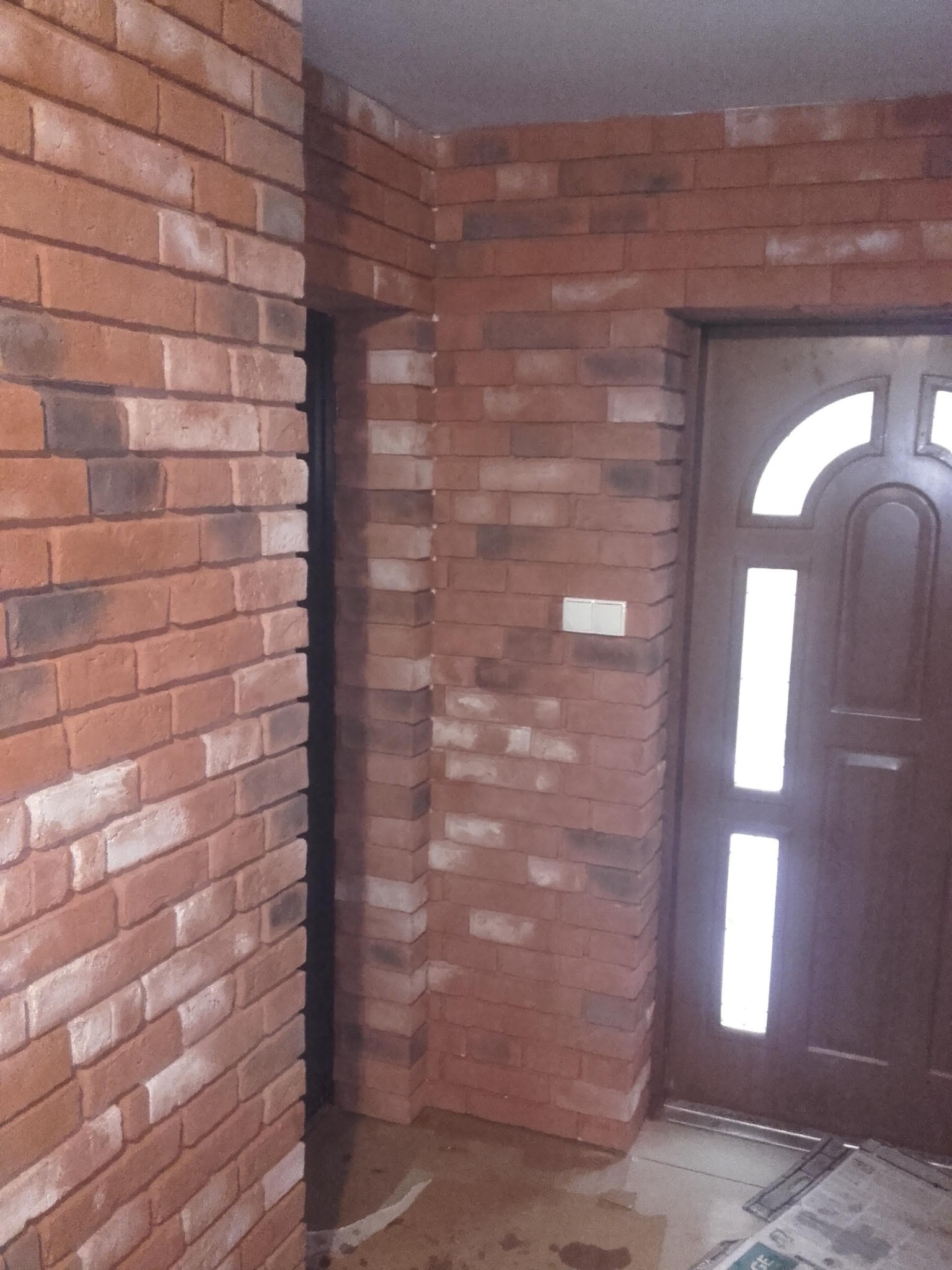 Dekoracyjna ściana z cegły klinkierowej w odcieniach czerwieni i brązu, obok brązowe drzwi z przeszkleniami. Widoczny fragment podłogi z drobnymi zabrudzeniami.