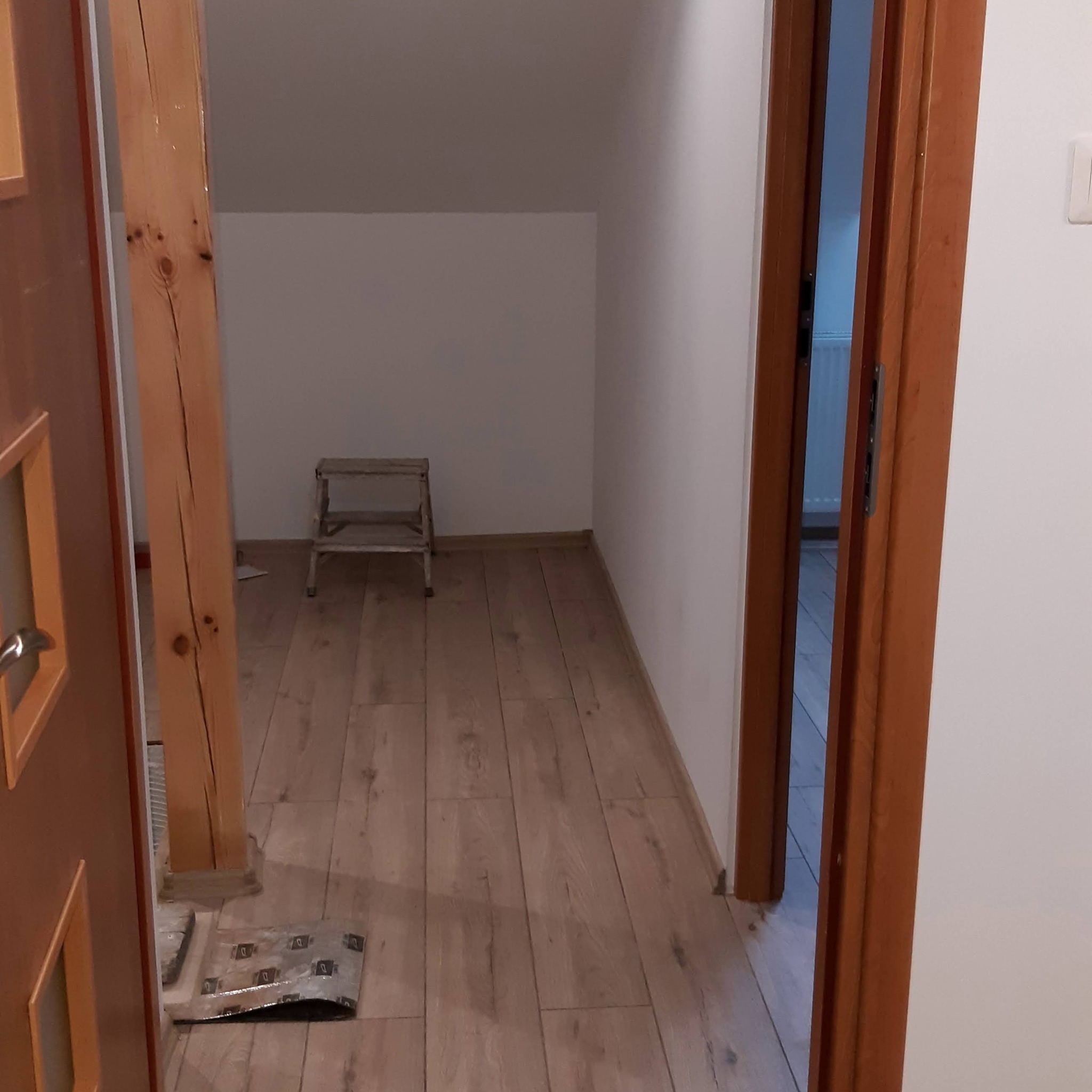 Poddasze z drewnianą podłogą i schodkami, widoczne przez otwarte drzwi. Ściany białe, w tle drewniany słup. Częściowo widoczne materiały budowlane.