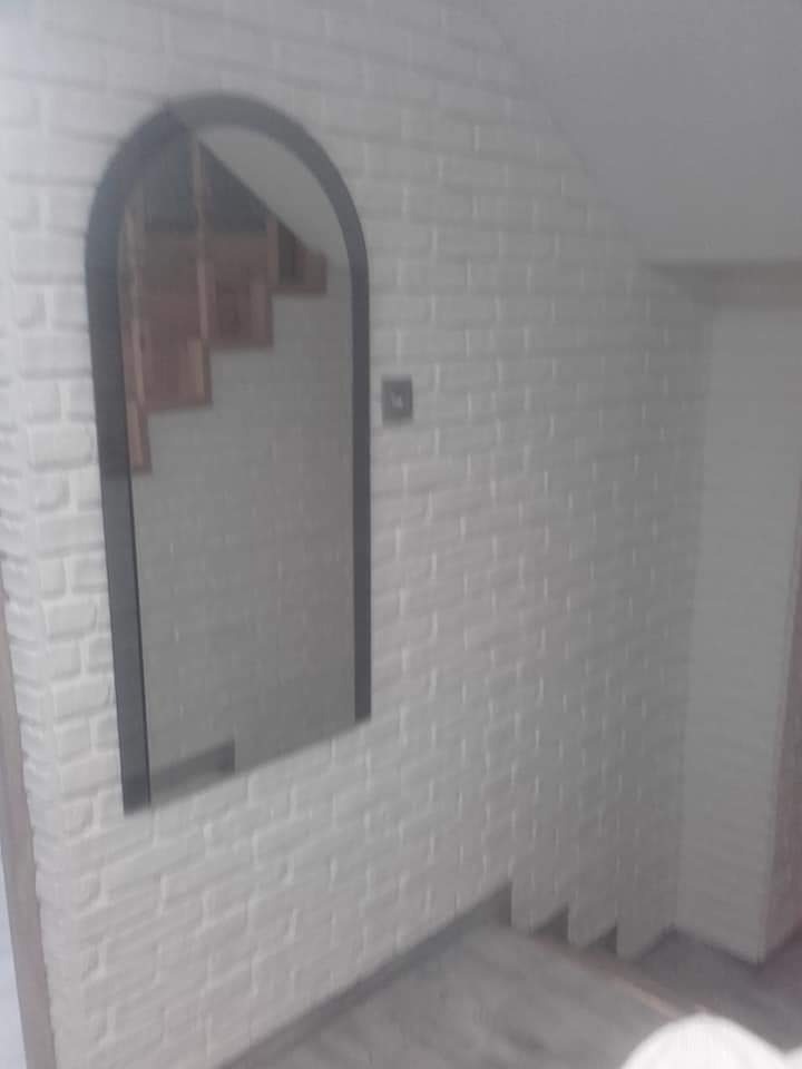 Biała ściana wykończona w stylu cegiełek z lustrem w czarnej ramie. Widoczne schody i fragment podłogi. Minimalistyczny design wnętrza.