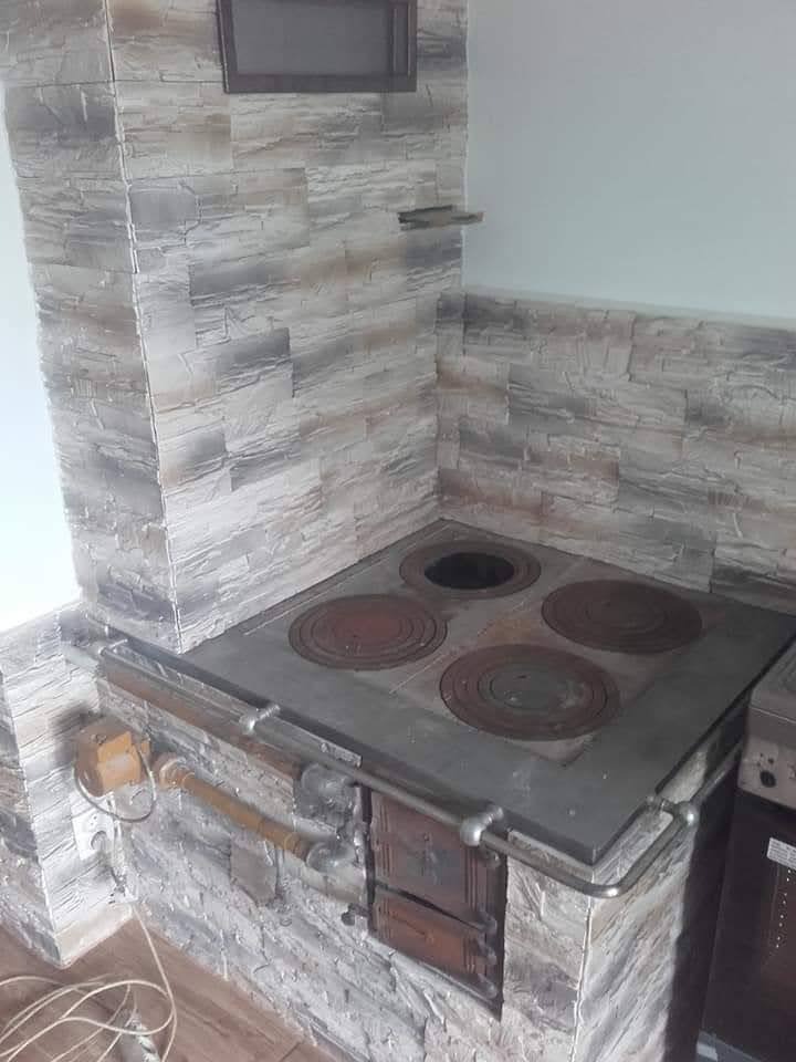 Wykończenie kuchni z piecem kaflowym obłożonym dekoracyjną, szarą cegłą. Widoczne palenisko z żeliwnymi płytami i fragment ściany z imitacją kamienia.