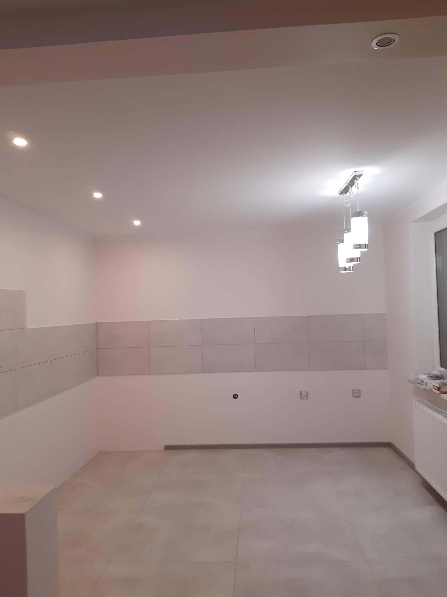 Jasne wnętrze kuchni po remoncie: szare płytki na ścianie i podłodze, nowoczesna lampa wisząca, gładkie ściany, widoczne gniazdka elektryczne. Minimalistyczny design.