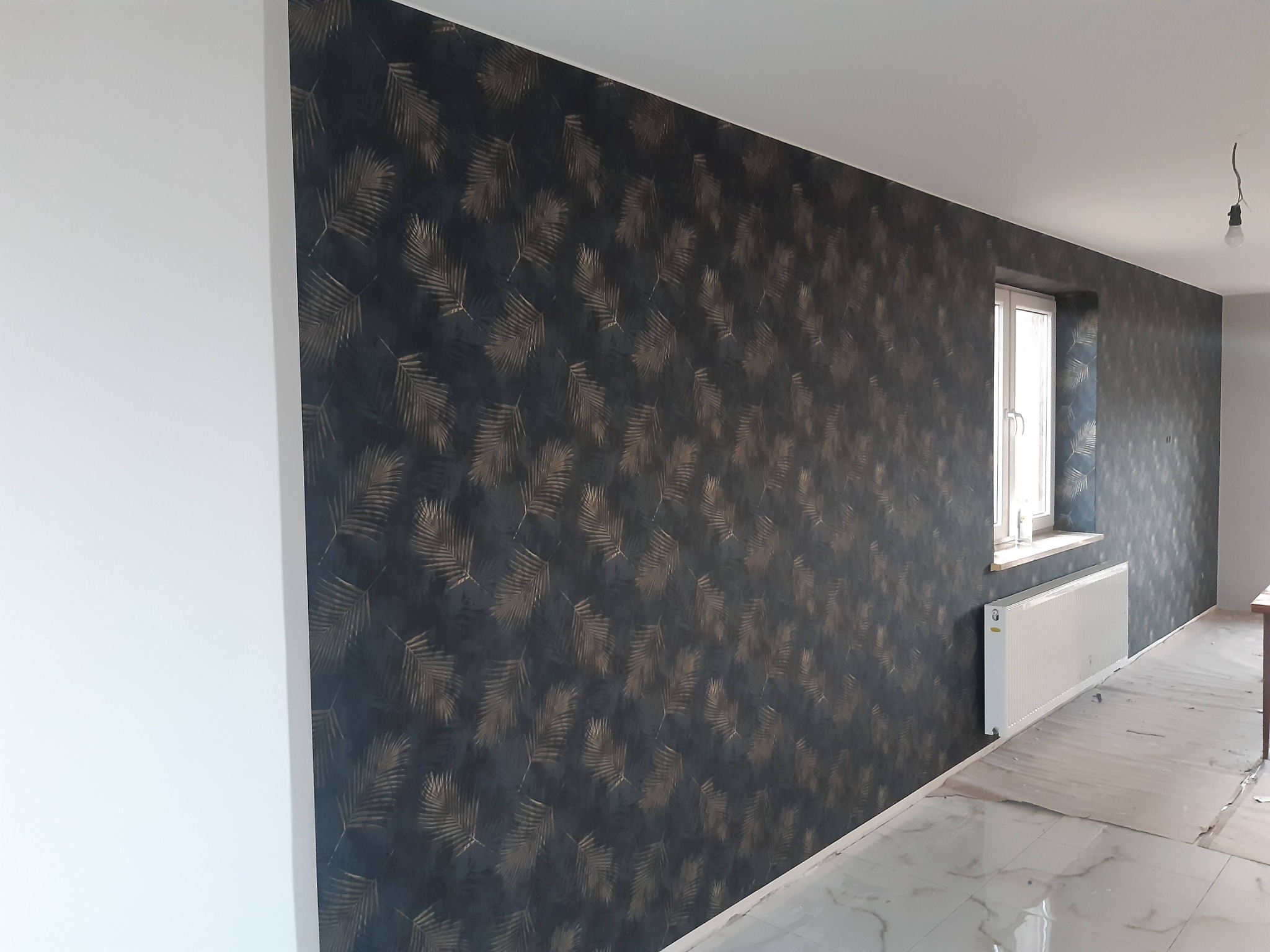 Ściana z ciemną tapetą w złote liście, biała ściana obok, okno z grzejnikiem. Podłoga wyłożona białymi płytkami imitującymi marmur. Wnętrze w trakcie wykańczania.