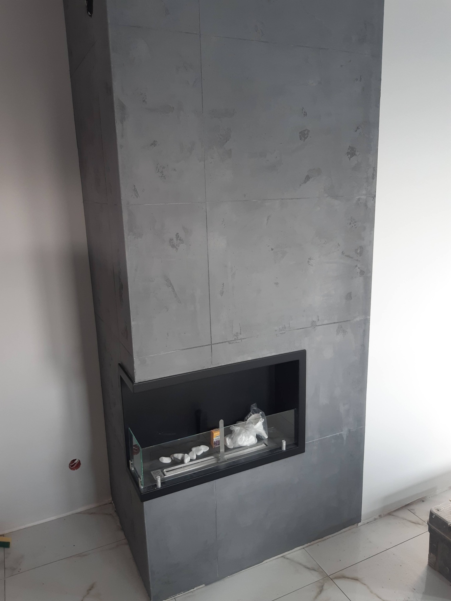 Nowoczesny biokominek obudowany szarą, betonową płytą dekoracyjną, wkomponowany w białą ścianę. Widoczne detale wykończenia i minimalistyczny design.
