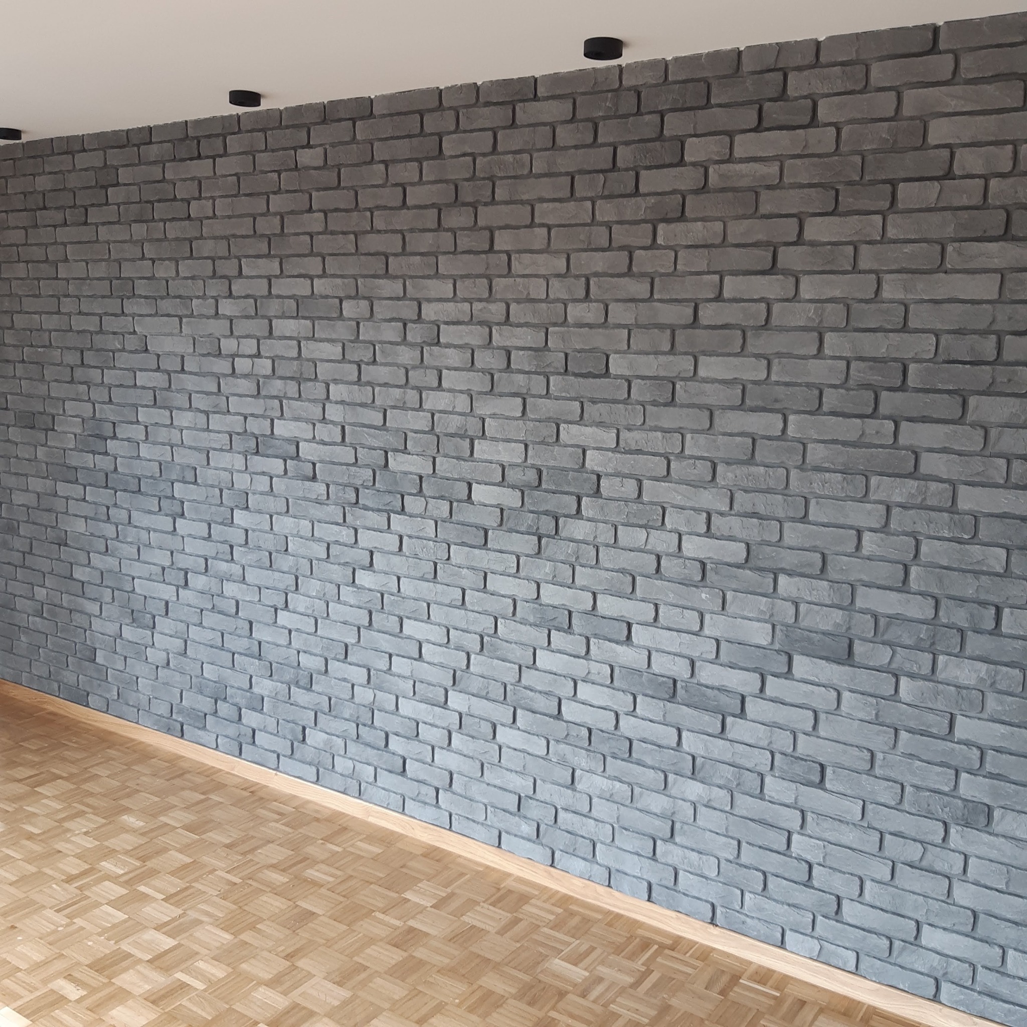 Szara ściana z cegły dekoracyjnej w nowym wnętrzu, oświetlona punktowo, z parkietem na podłodze. Widok na fragment pomieszczenia po wykończeniu.
