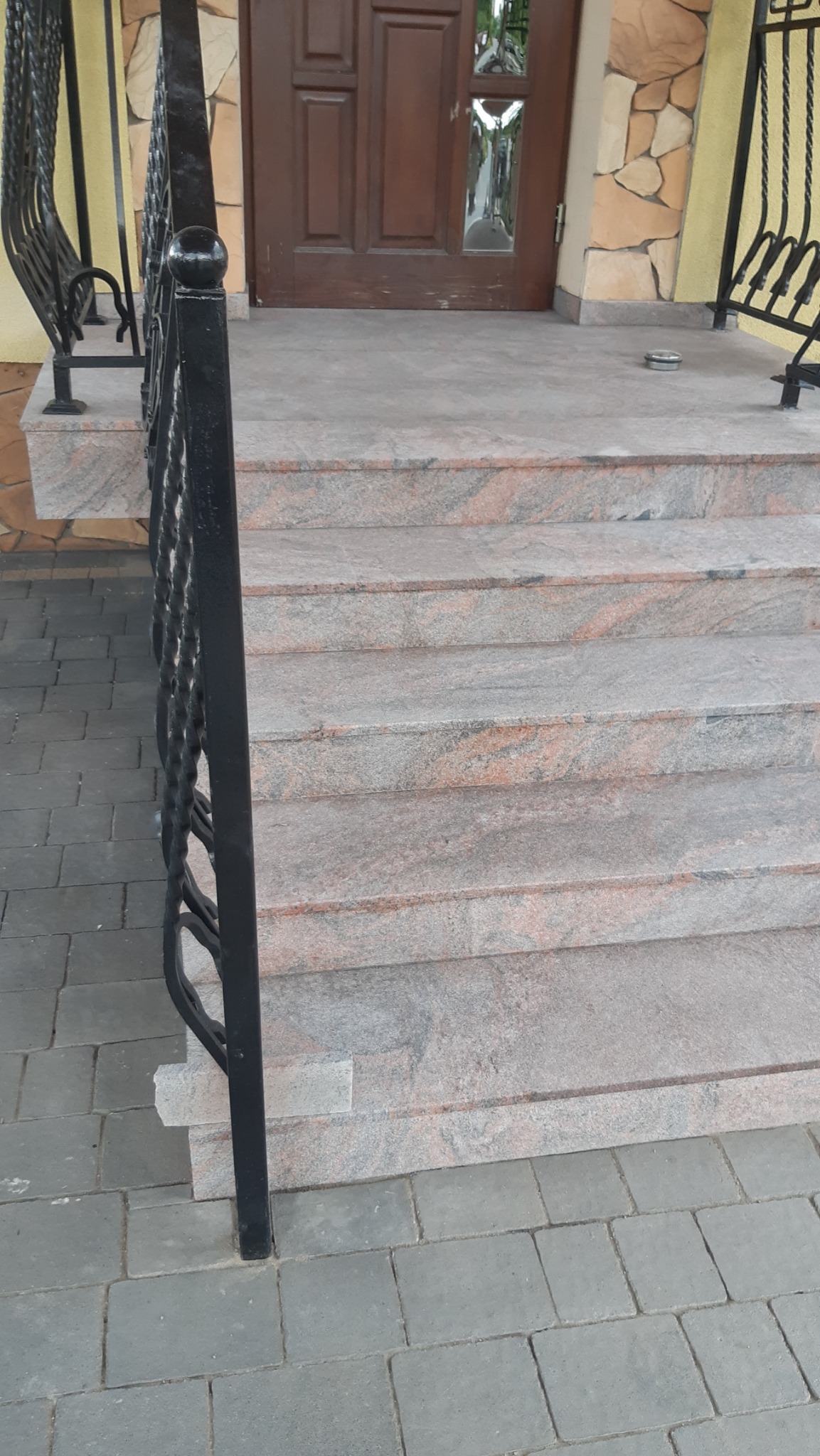 Granitowe schody wejściowe z kutą balustradą, prowadzące do brązowych drzwi. Kamienna elewacja budynku w tle. Widok z dołu, podkreślający solidność wykonania.