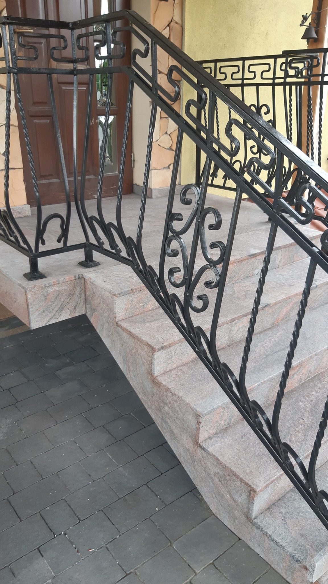Schody wejściowe z jasnego kamienia z czarną, kutą balustradą o zdobnym wzorze. Widoczna kostka brukowa i fragment drzwi wejściowych. Ujęcie z dołu.