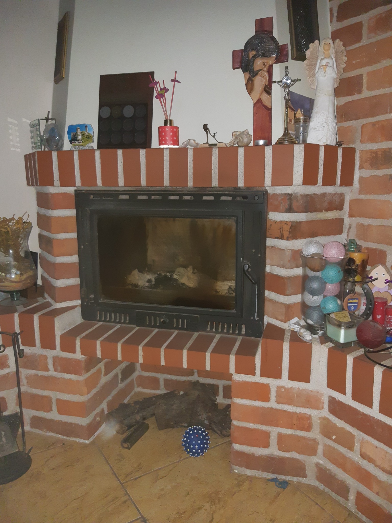 Murowany kominek z czarnym wkładem, na półce dekoracje: figurki, świeczki, pamiątki. W palenisku drewno, obok niebieska piłka. Ciepłe oświetlenie.