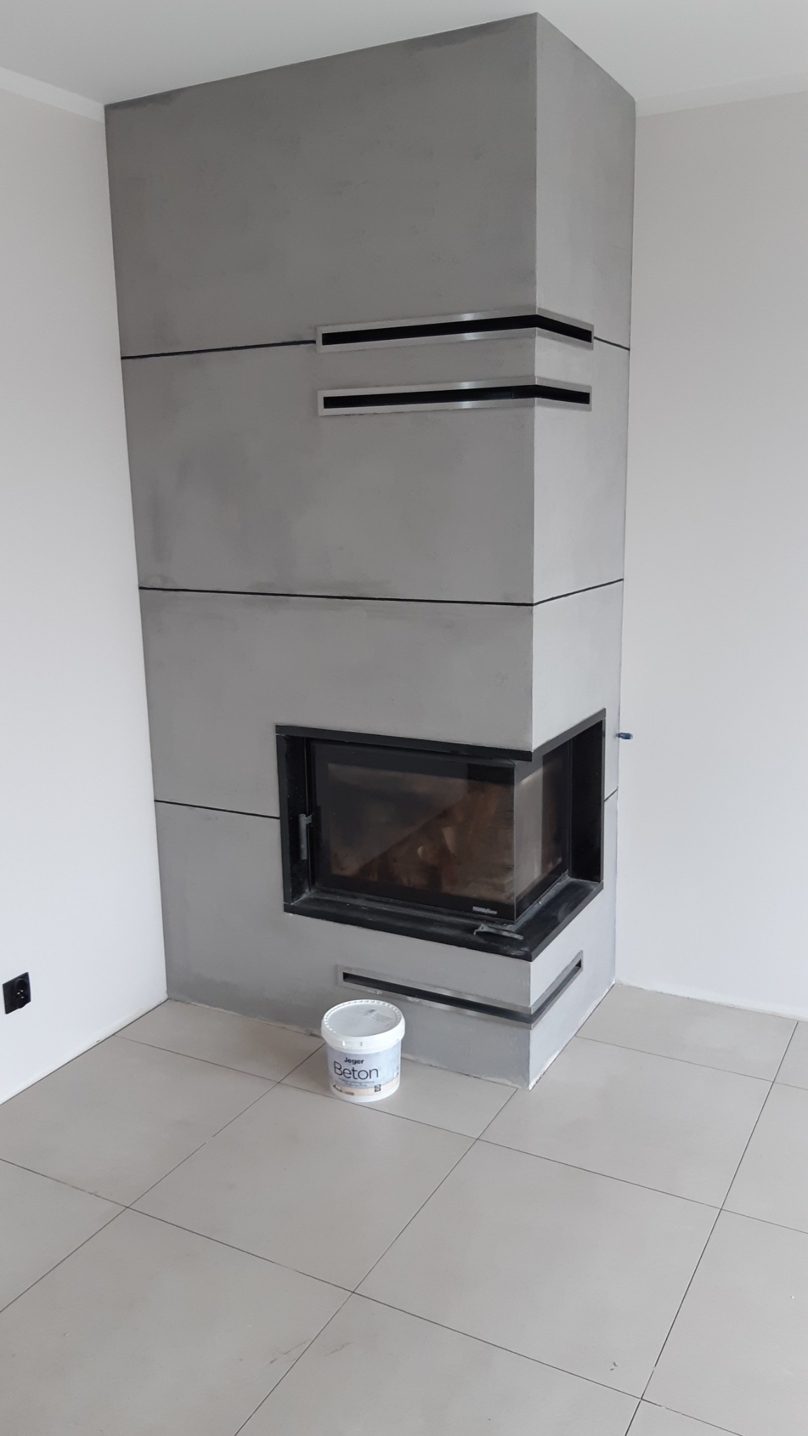 Wykończony kominek narożny w szarym betonie architektonicznym z czarnym wkładem, na podłodze z płytek. Wiadro z farbą 'Beton' stoi obok. Minimalistyczny design.