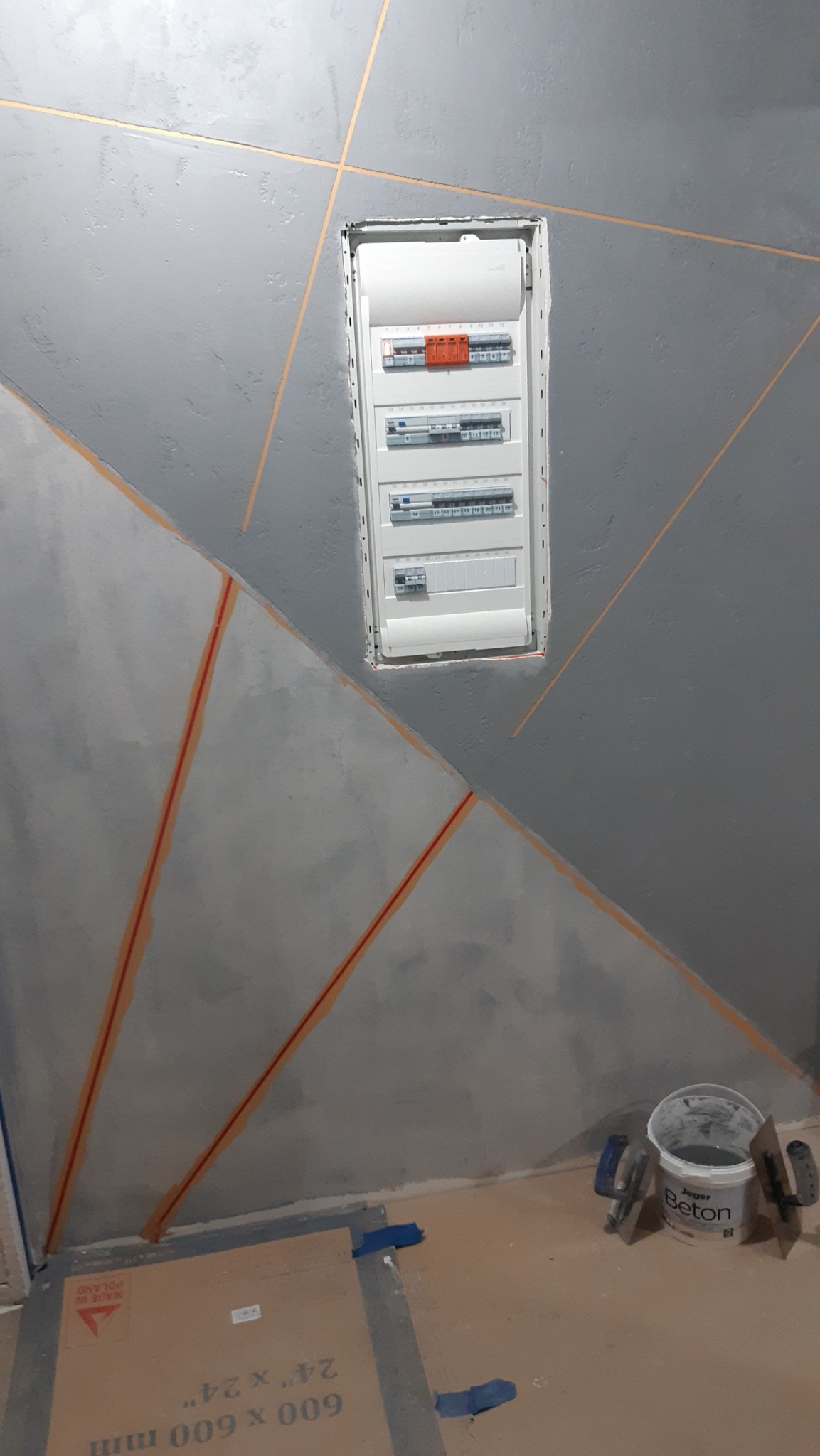 Ściana z szarą, dekoracyjną strukturą i geometrycznymi, pomarańczowymi liniami. W ścianie wnęka z białą skrzynką elektryczną. Widoczne narzędzia i wiadro z napisem Beton.