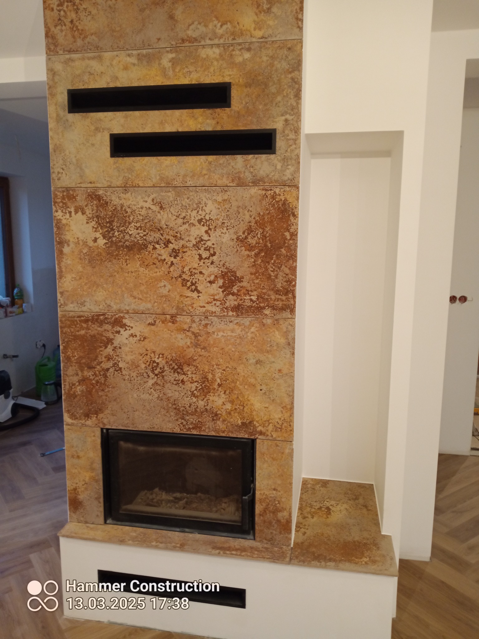 Obudowa kominka z rustykalnych, brązowo-beżowych płyt, z czarnymi wnękami wentylacyjnymi i wkładem kominkowym. Obok wnęka w białej ścianie. Parkiet w jodełkę na podłodze.