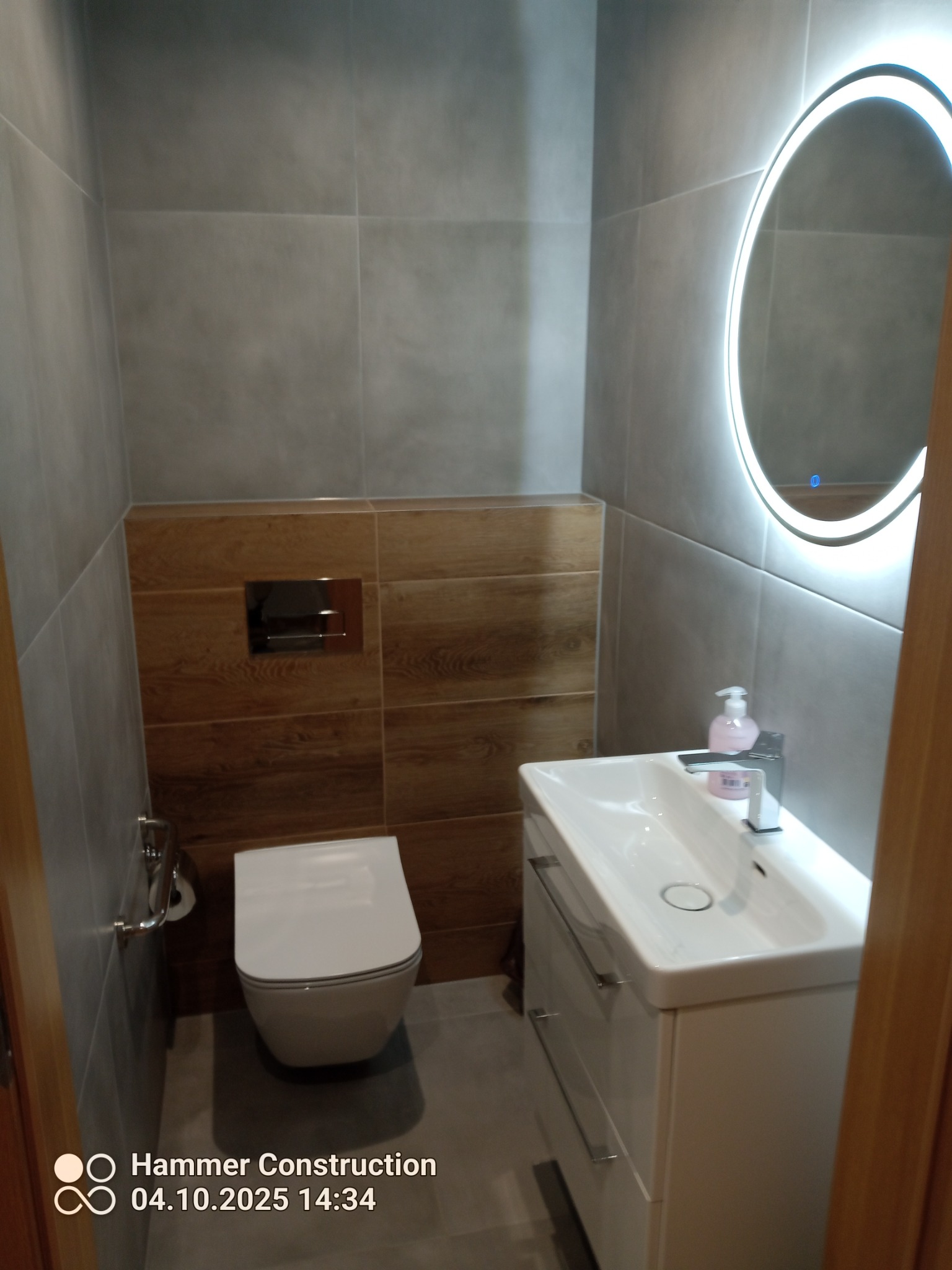 Nowoczesna łazienka po remoncie: szare płytki, drewnopodobne akcenty, podświetlane lustro owalne, biała umywalka z szafką i wisząca toaleta. Minimalistyczny design.