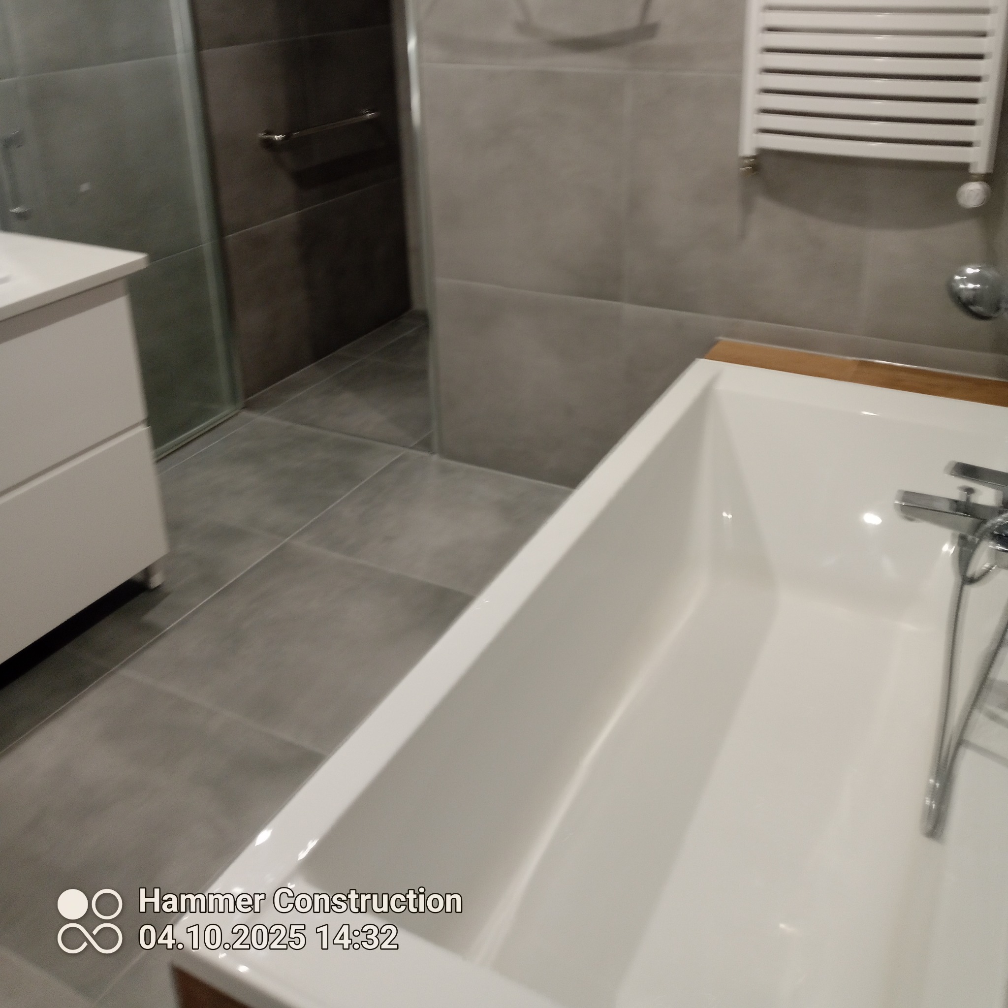 Nowoczesna łazienka z wanną, szarą glazurą i białymi meblami. Widoczny grzejnik drabinkowy i szklana kabina prysznicowa. Minimalistyczny design.