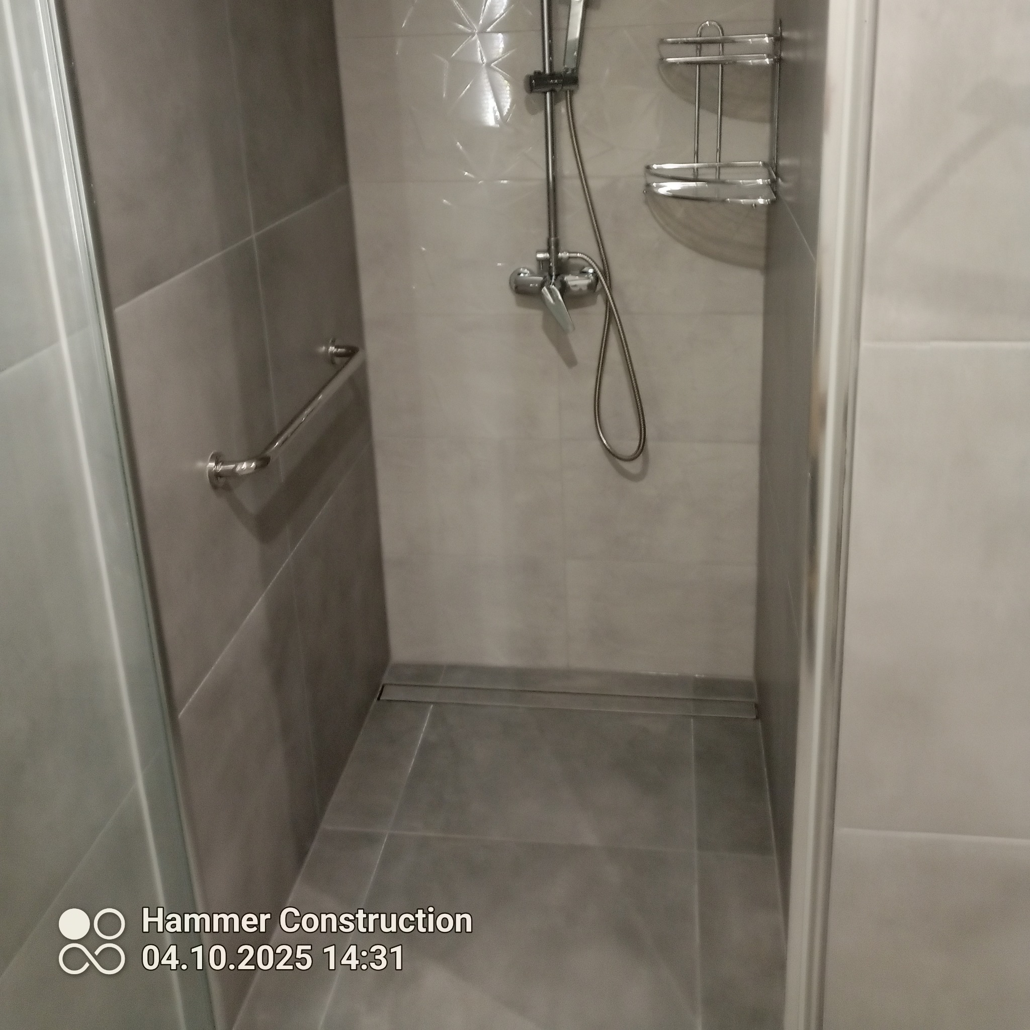 Szara kabina prysznicowa z odpływem liniowym, chromowaną armaturą i półką. Ściany wyłożone płytkami w odcieniach szarości. Widoczny uchwyt dla niepełnosprawnych.