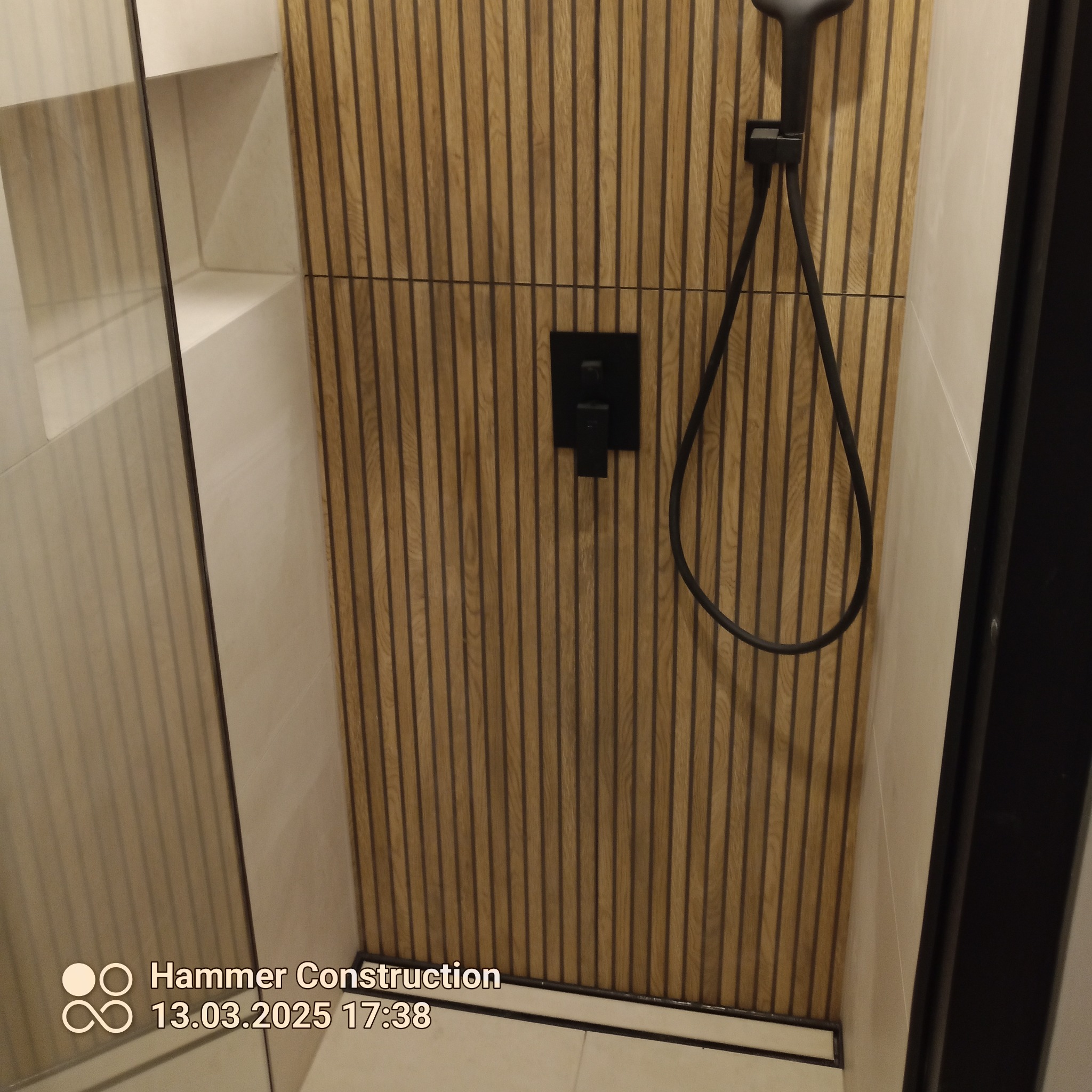 Nowoczesna kabina prysznicowa z drewnianą ścianą, czarną armaturą i odpływem liniowym. Minimalistyczny design, jasne płytki, eleganckie wykończenie.