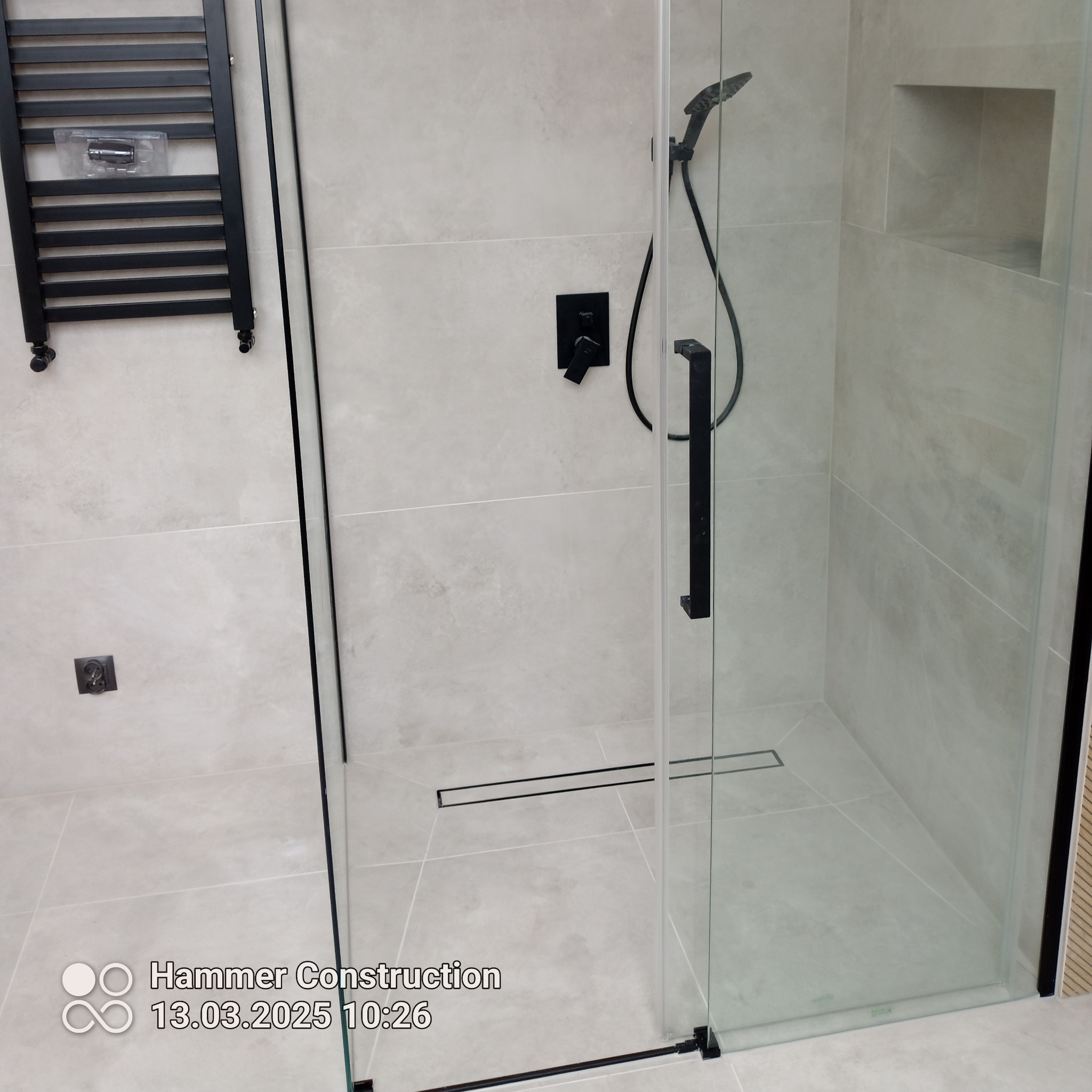 Nowoczesna kabina prysznicowa z czarnymi elementami: armatura, grzejnik drabinkowy, odpływ liniowy. Jasne płytki, wnęka w ścianie. Minimalistyczny design.