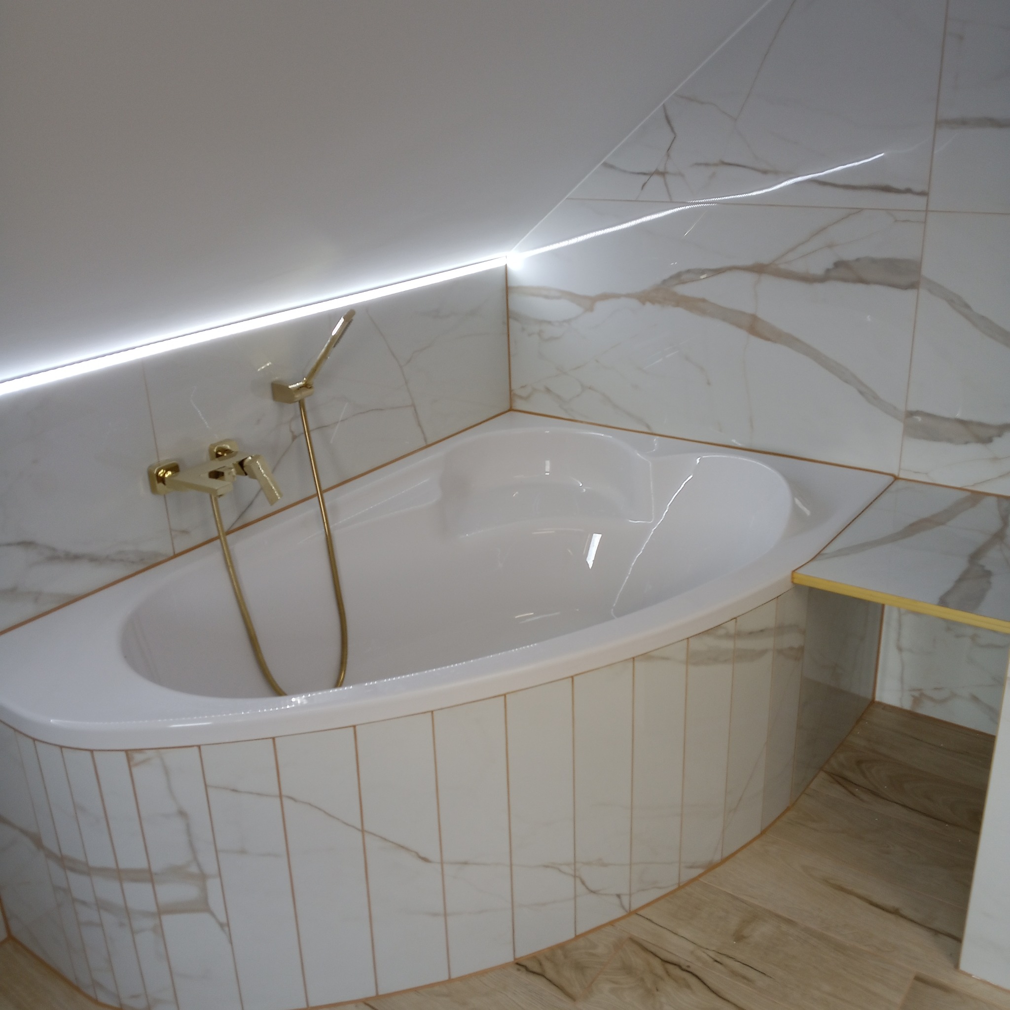 Biała wanna narożna z armaturą w kolorze złota, obudowana płytkami imitującymi marmur. Podświetlenie LED nad wanną. Ukośny sufit. Wykończenie łazienki w stylu glamour.