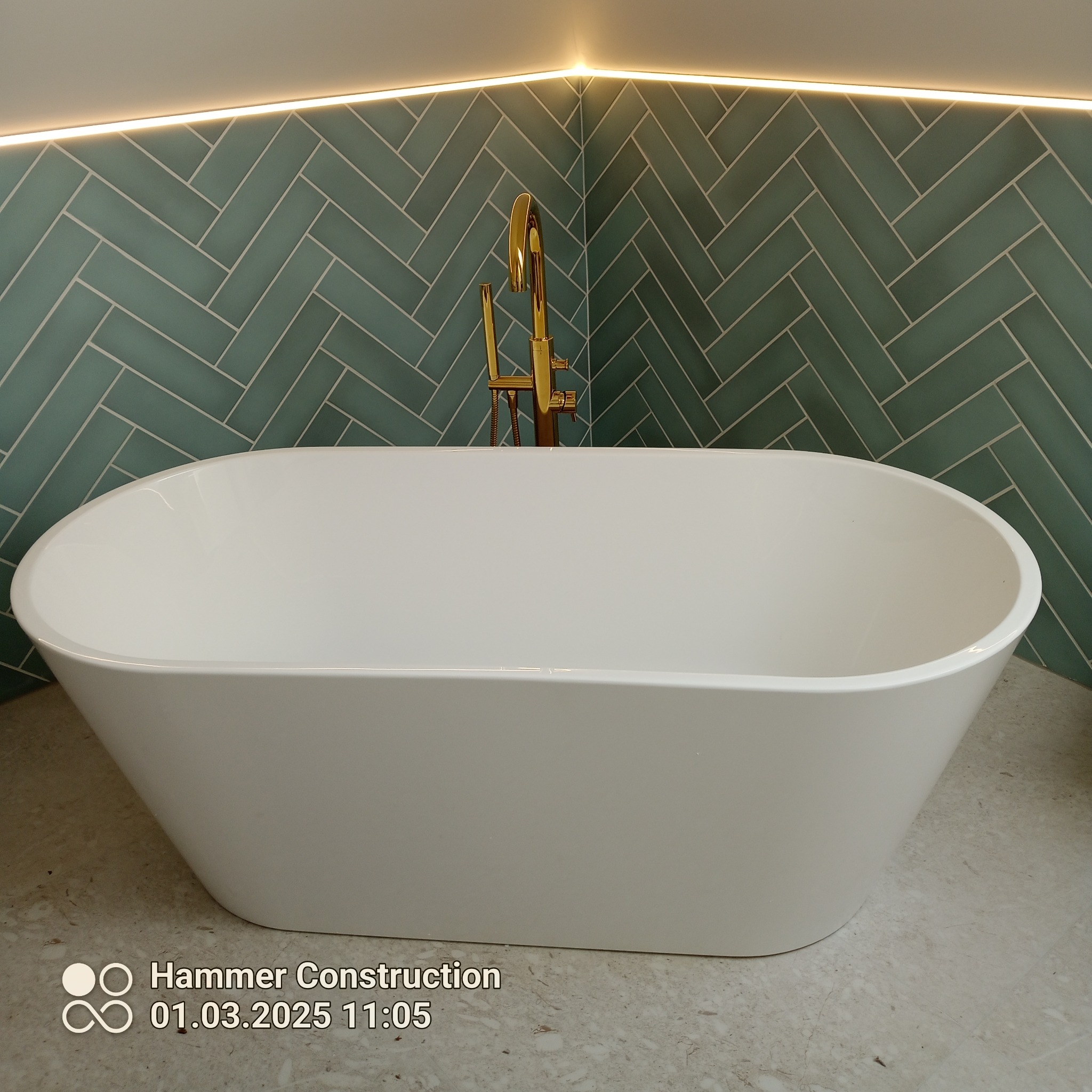 Biała wanna wolnostojąca z złotą armaturą na tle ściany wyłożonej zieloną, ułożoną w jodełkę glazurą, oświetloną ledowym światłem. Nowoczesna aranżacja łazienki.