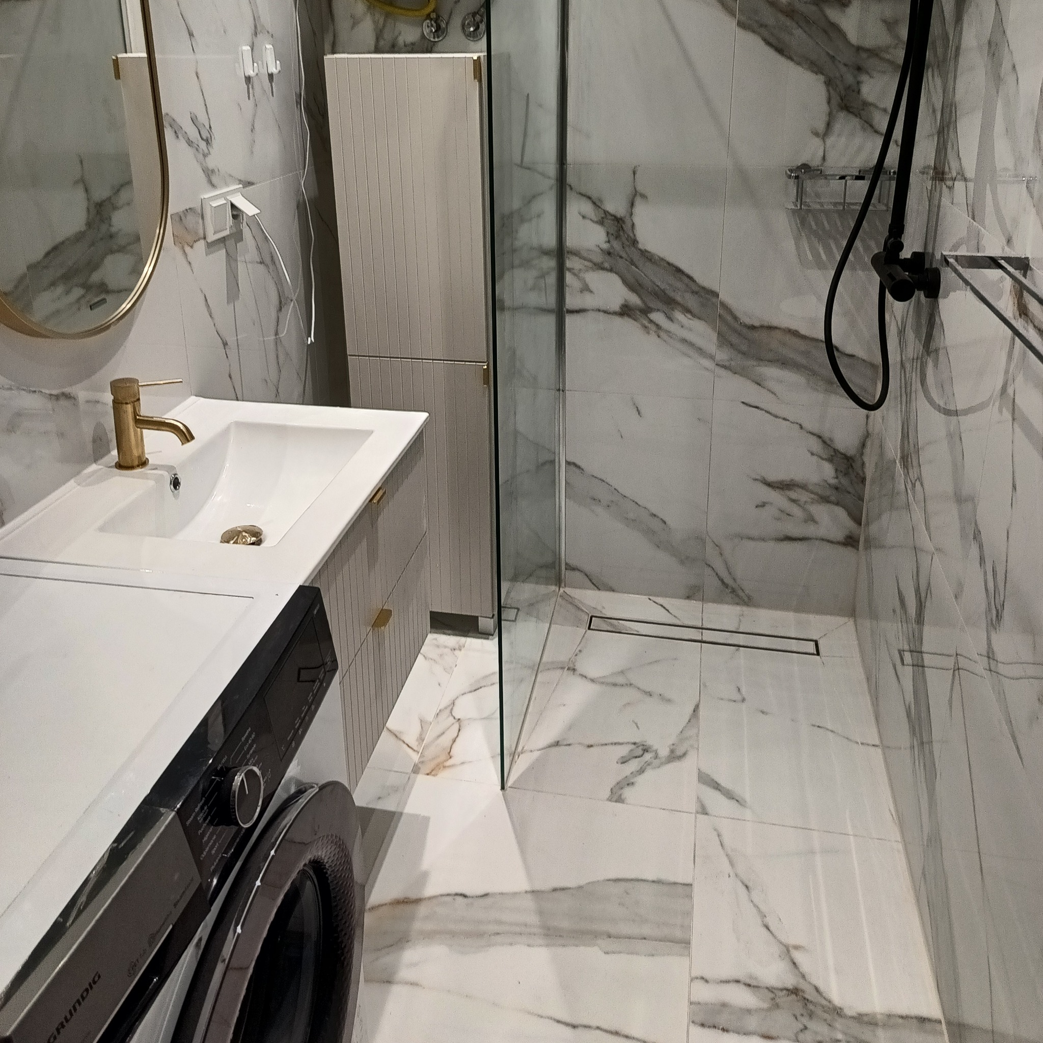 Nowoczesna łazienka z marmurowymi płytkami, kabiną prysznicową ze szkła, umywalką z złotą armaturą i pralką. Minimalistyczny design, jasne wnętrze.