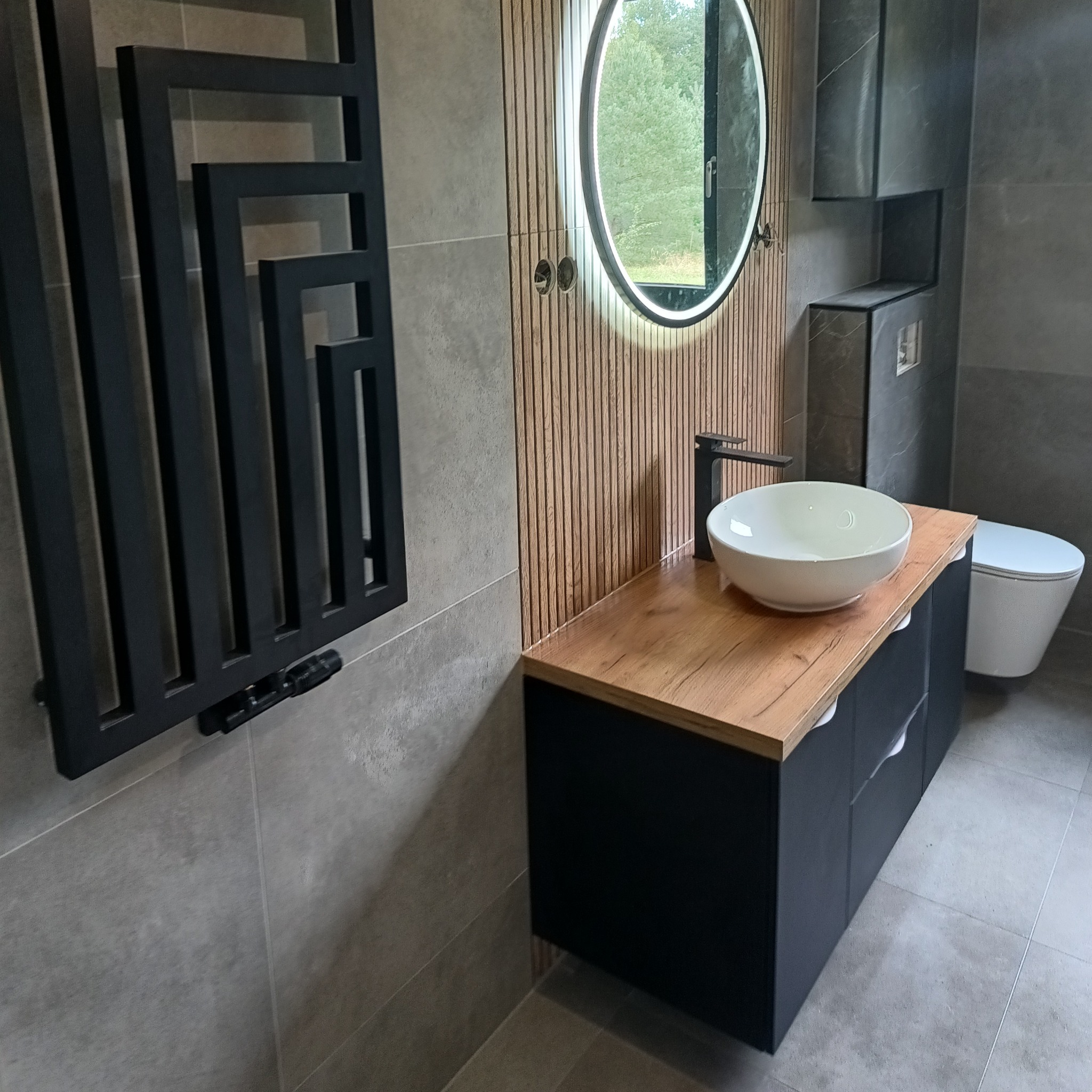 Nowoczesna łazienka z okrągłym lustrem LED, drewnianym blatem, białą umywalką i czarnym grzejnikiem drabinkowym na szarej płytce. Minimalistyczny design.