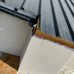 Paweł Drej Dachy - Szary dach z blachy trapezowej z rynną w narożniku budynku, widok z dołu, Kielce. Widoczne detale obróbki blacharskiej i struktura elewacji.