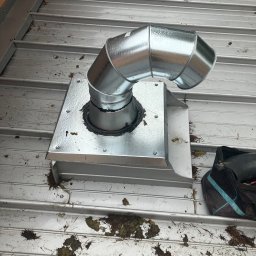 Paweł Drej Dachy - Metalowy komin wentylacyjny na dachu z blachy trapezowej, uszczelniony czarną gumą. Widoczne ślady liści i zabrudzeń na powierzchni dachu. Ujęcie z góry.