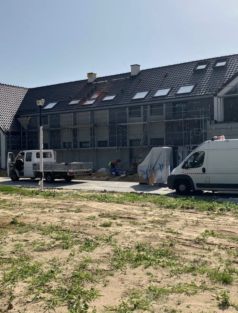 Budynek w trakcie ocieplania, widoczne rusztowania i materiały izolacyjne. Na pierwszym planie dwa samochody dostawcze i pracownicy w trakcie pracy.