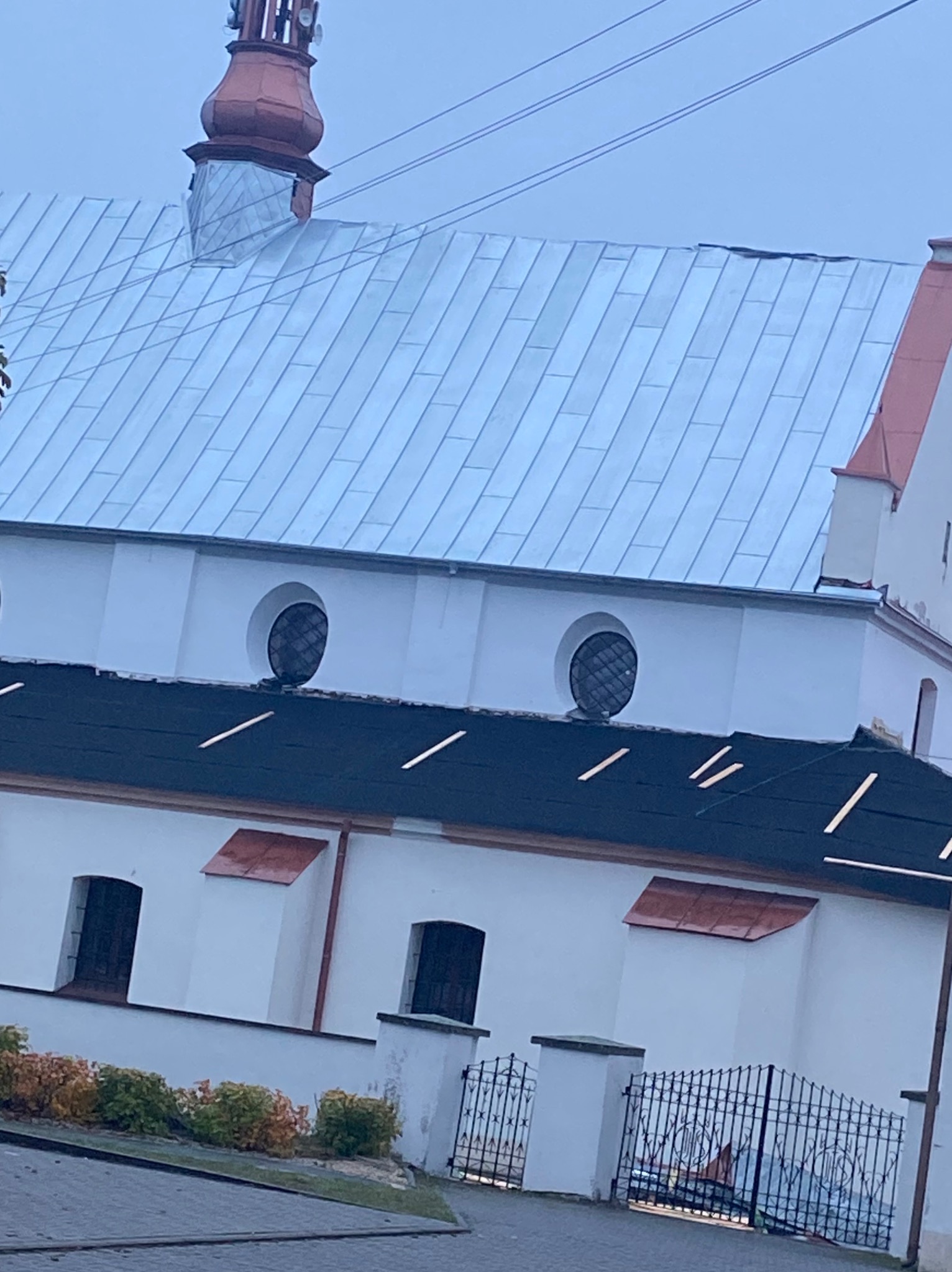 Biały kościół z metalowym dachem w odcieniach szarości, widoczny z dołu pod kątem. Na dachu wieżyczka z czerwonym dachem. W tle pochmurne niebo.