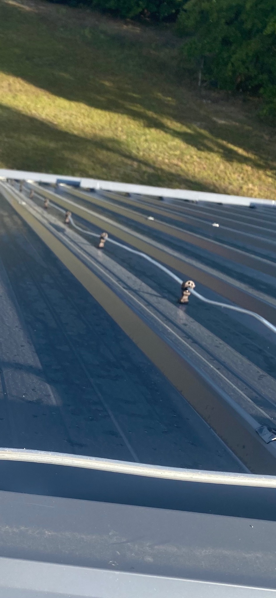 Szary dach z blachy trapezowej z zamontowanymi uchwytami do instalacji odgromowej i widocznym fragmentem rynny. Ujęcie z dołu, w tle zieleń.