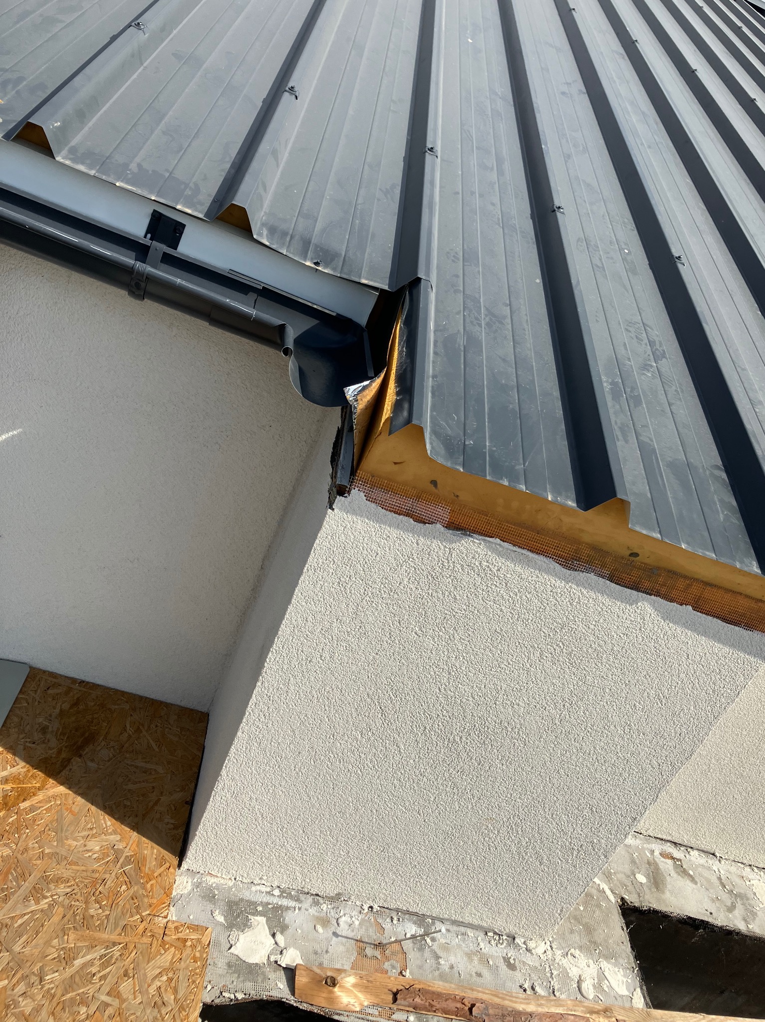 Szary dach z blachy trapezowej z rynną w narożniku budynku, widok z dołu, Kielce. Widoczne detale obróbki blacharskiej i struktura elewacji.