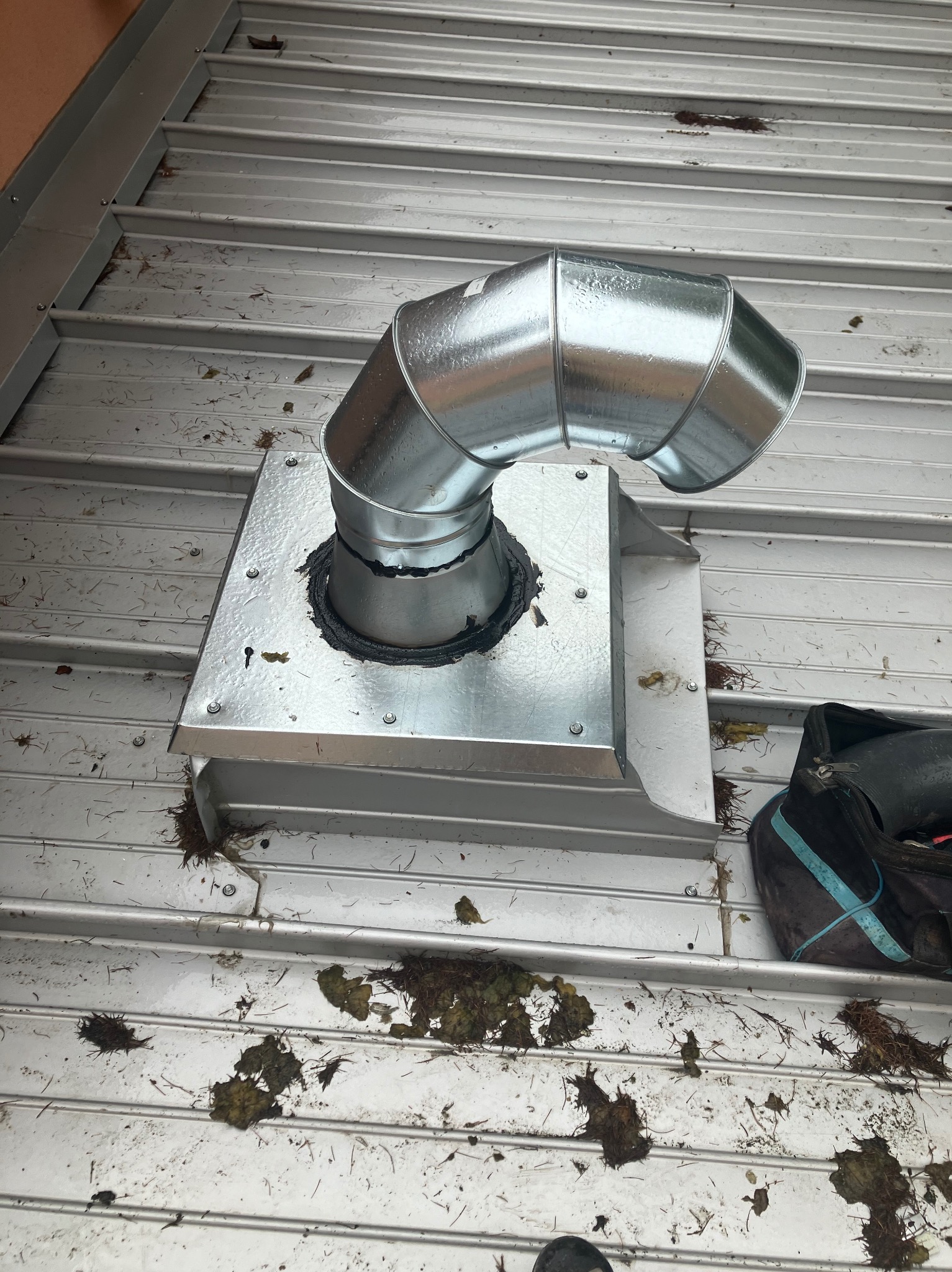 Metalowy komin wentylacyjny na dachu z blachy trapezowej, uszczelniony czarną gumą. Widoczne ślady liści i zabrudzeń na powierzchni dachu. Ujęcie z góry.