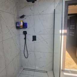 Paweł Gębala - Kabina prysznicowa walk-in z czarną armaturą i odpływem liniowym, wykończona płytkami imitującymi marmur. Na ścianie półka z kosmetykami.