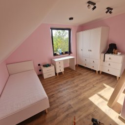 Paweł Gębala - Pokój na poddaszu z białymi meblami: łóżko, biurko, komoda i szafa. Na podłodze narzędzia. Ściany pomalowane na różowo. Widok na zewnątrz przez okno.
