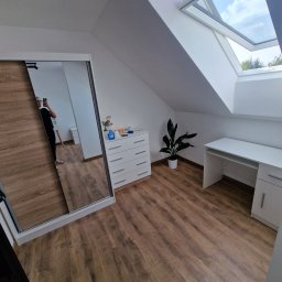 Paweł Gębala - Jasne poddasze z drewnianą podłogą, białymi meblami i oknem dachowym. W lustrze szafy odbija się osoba robiąca zdjęcie. Minimalistyczny wystrój.