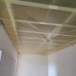 Przygotowanie sufitu do prac wykończeniowych: drewniana konstrukcja zabezpieczona folią i taśmą malarską w rogu pomieszczenia z fragmentem ściany z płyty gipsowej.