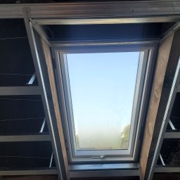 Montaż okna dachowego w konstrukcji metalowej, widok od wewnątrz. Widoczne elementy mocujące i fragment nieba przez szybę. Drewniane elementy wykończeniowe.