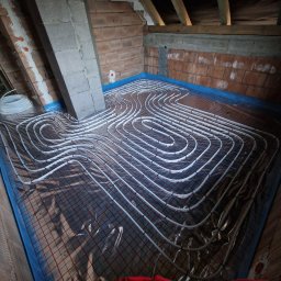 Białe rury ogrzewania podłogowego ułożone na folii z siatką w pomieszczeniu z ceglanymi ścianami i drewnianą konstrukcją dachu. Widok z góry na instalację przed wylaniem posadzki.