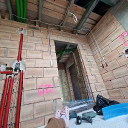 Paweł Gębala - Surowy stan wnętrza z instalacjami: czerwone rury, zielone peszle, białe kable. Widoczne narzędzia i fragment osoby w różowych butach. Ściany z cegły dziurawki.