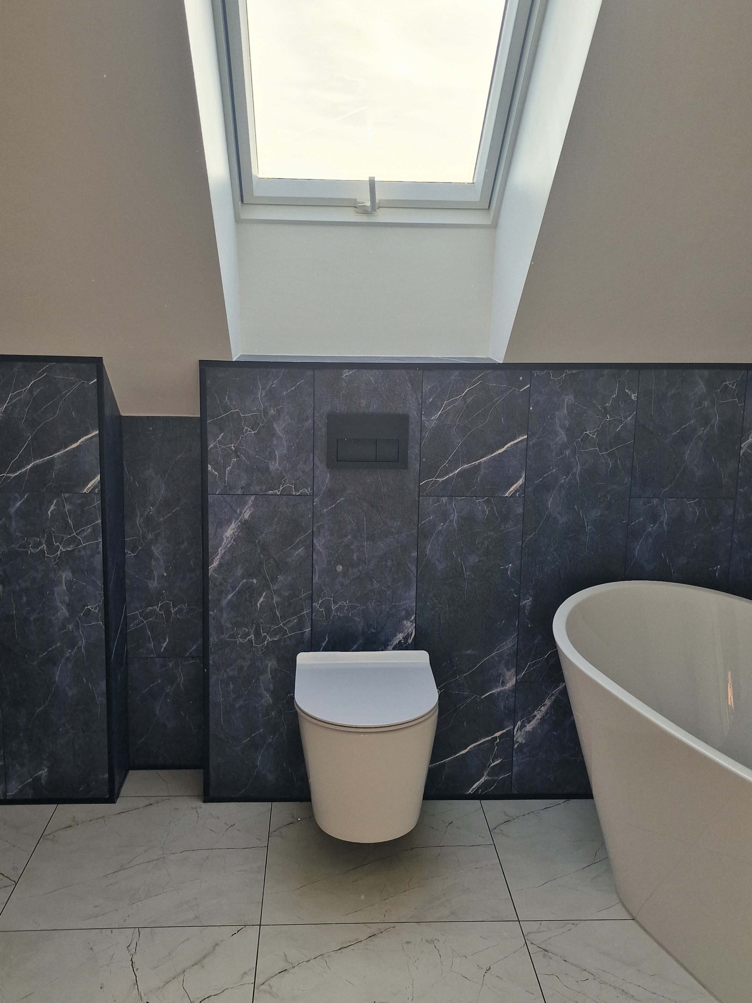 Wykończona łazienka na poddaszu z oknem dachowym. Ciemne płytki imitujące marmur na ścianach, biała wolnostojąca wanna i toaleta. Podłoga wyłożona jasnymi płytkami.