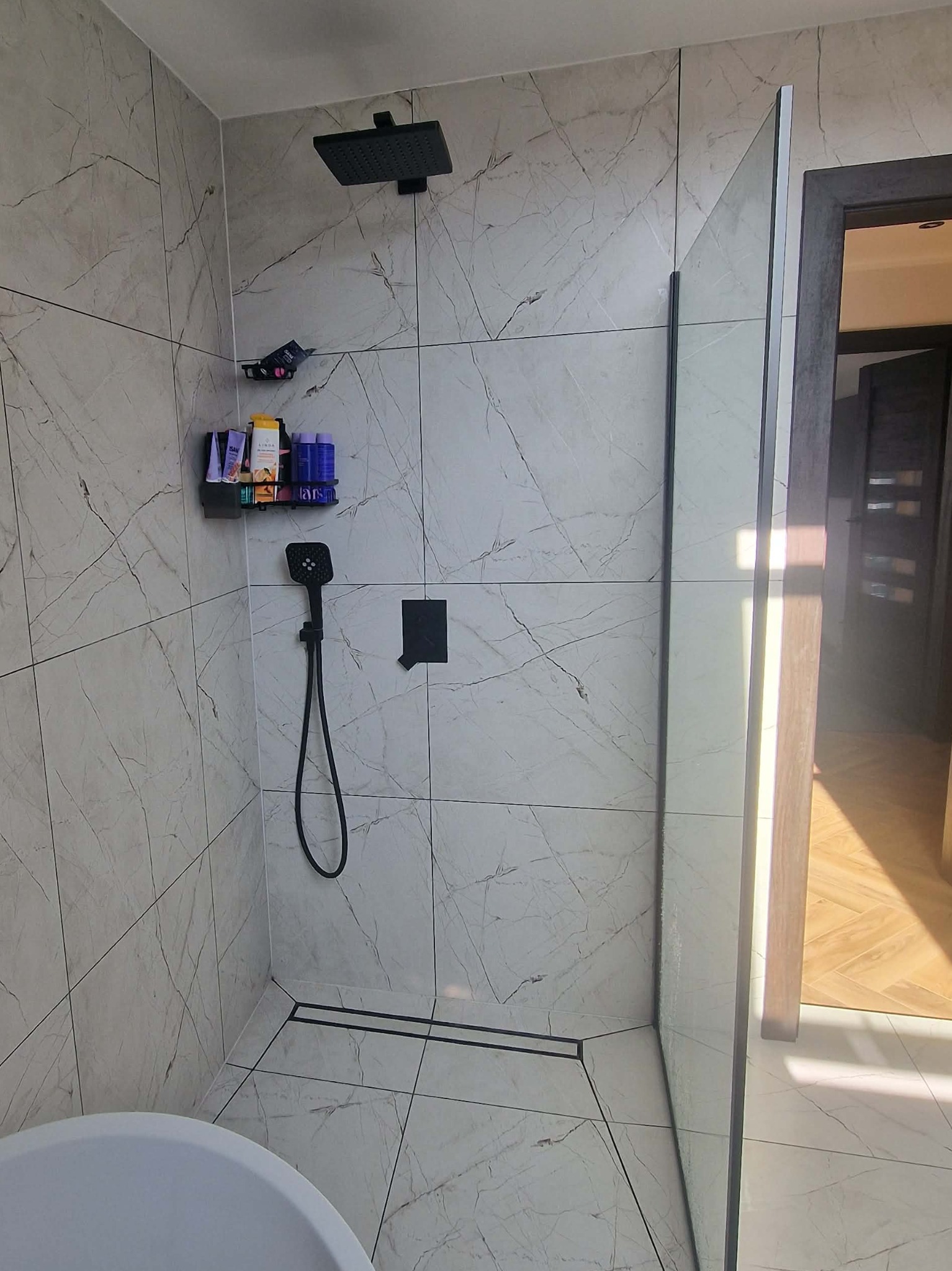 Kabina prysznicowa walk-in z czarną armaturą i odpływem liniowym, wykończona płytkami imitującymi marmur. Na ścianie półka z kosmetykami.