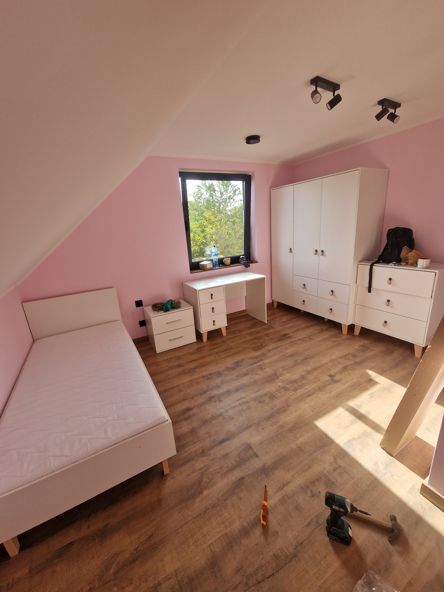 Pokój na poddaszu z białymi meblami: łóżko, biurko, komoda i szafa. Na podłodze narzędzia. Ściany pomalowane na różowo. Widok na zewnątrz przez okno.