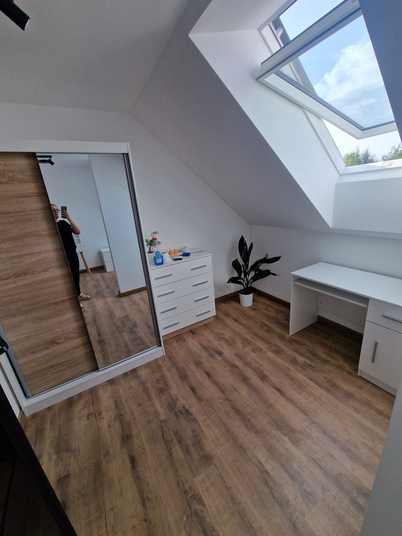Jasne poddasze z drewnianą podłogą, białymi meblami i oknem dachowym. W lustrze szafy odbija się osoba robiąca zdjęcie. Minimalistyczny wystrój.