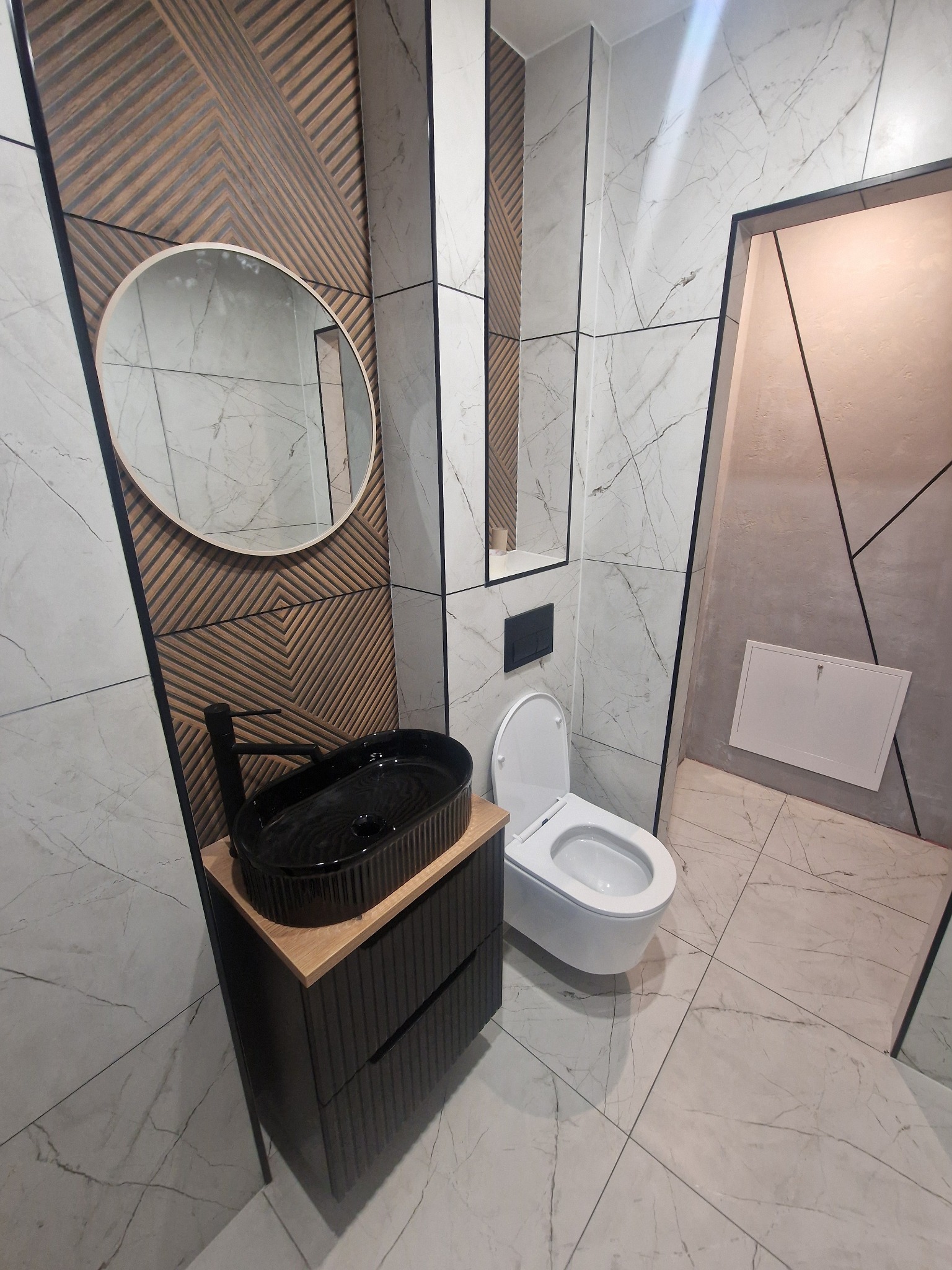 Nowoczesna łazienka z umywalką nablatową, okrągłym lustrem i marmurowymi płytkami. Dekoracyjna ściana z drewnianymi panelami dodaje unikalnego charakteru.