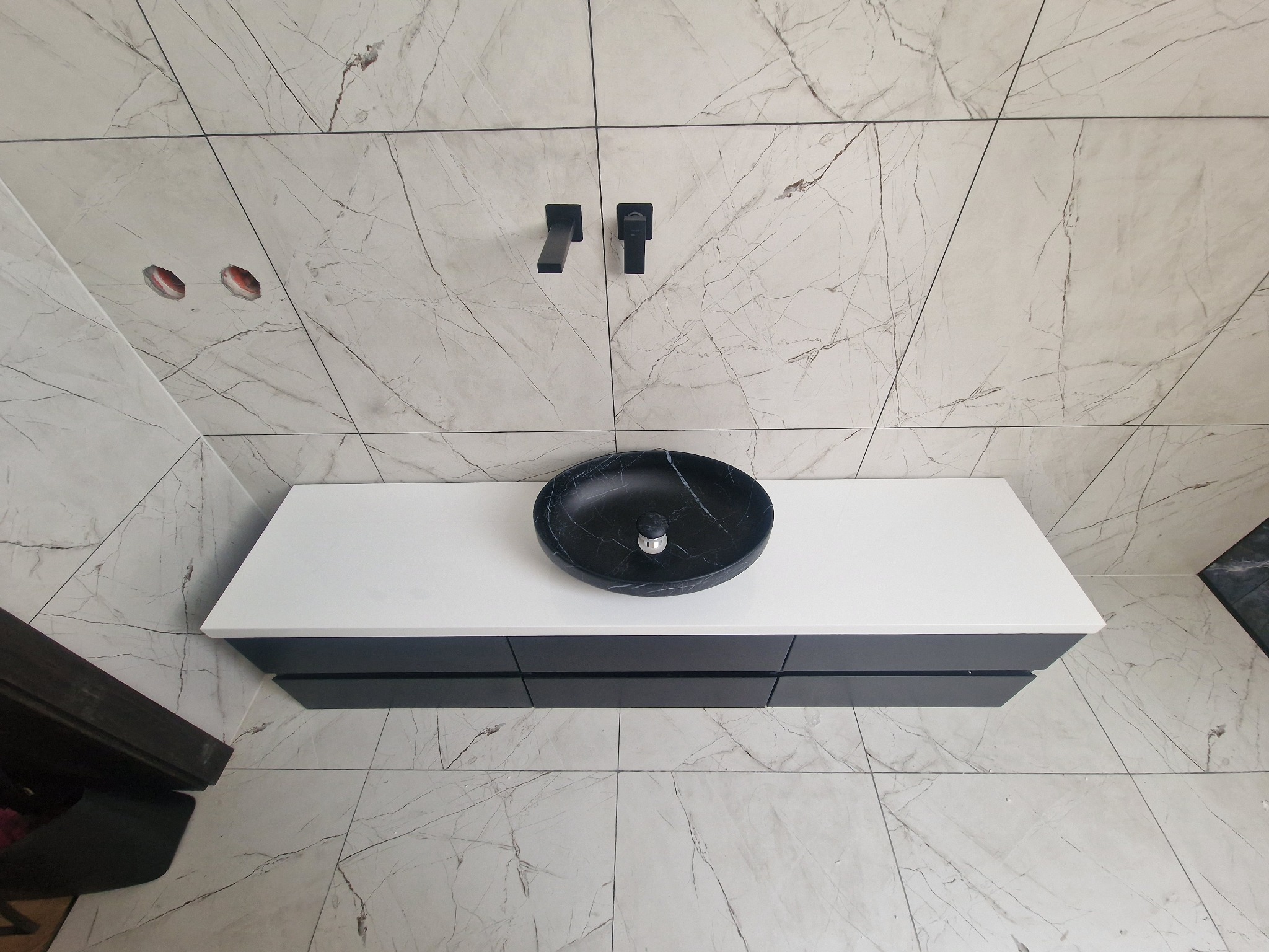 Nowoczesna łazienka z marmurowymi płytkami, czarną umywalką nablatową i minimalistyczną armaturą. Widoczne niedokończone otwory na ścianie.